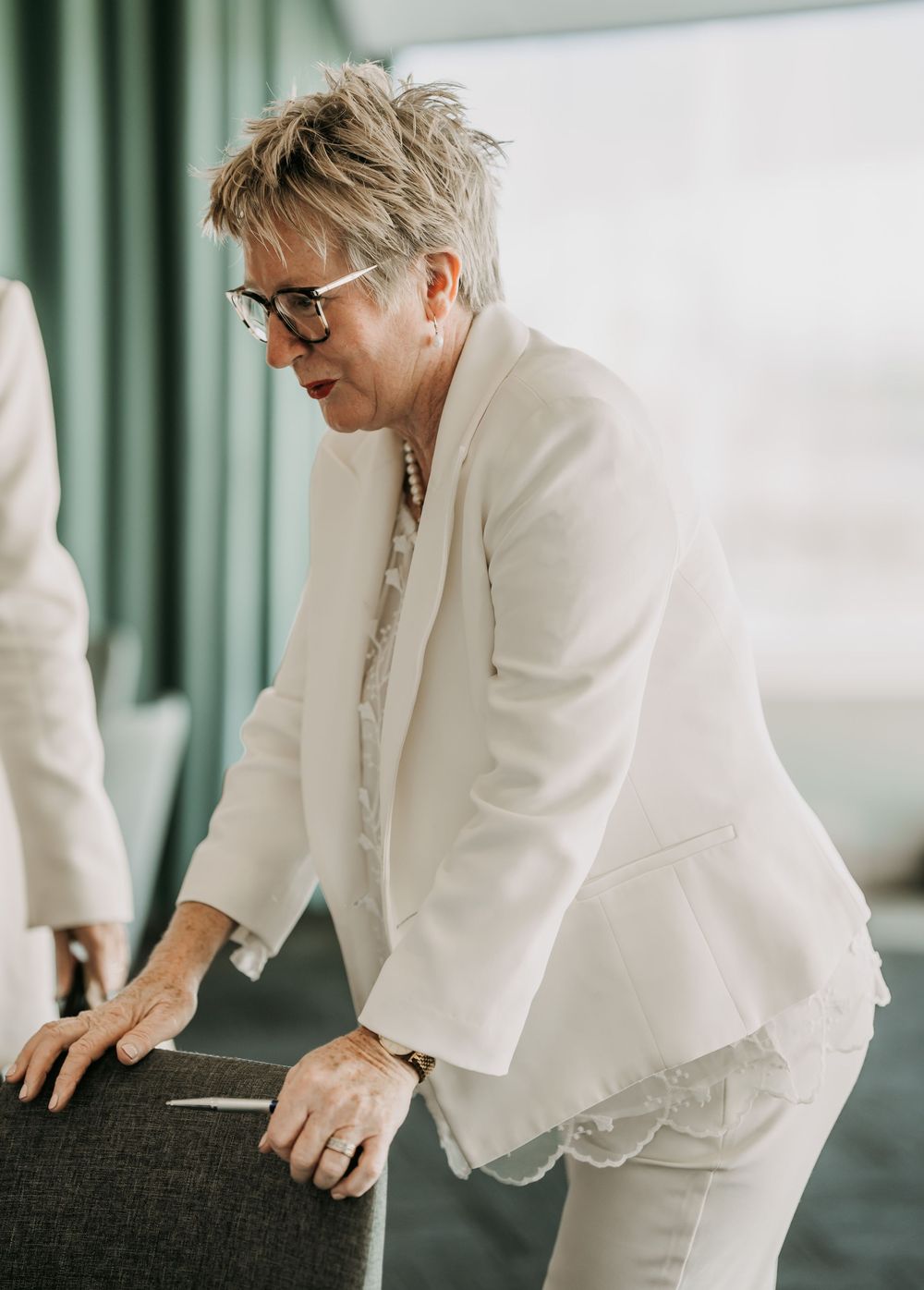 Person in a white blazer and glasses leaning against a dark surface in an indoor setting.