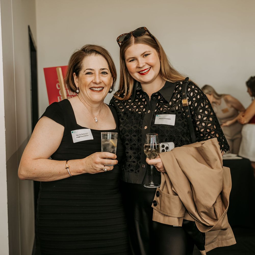 Two women hold drinks and smile while attending an indoor social event.