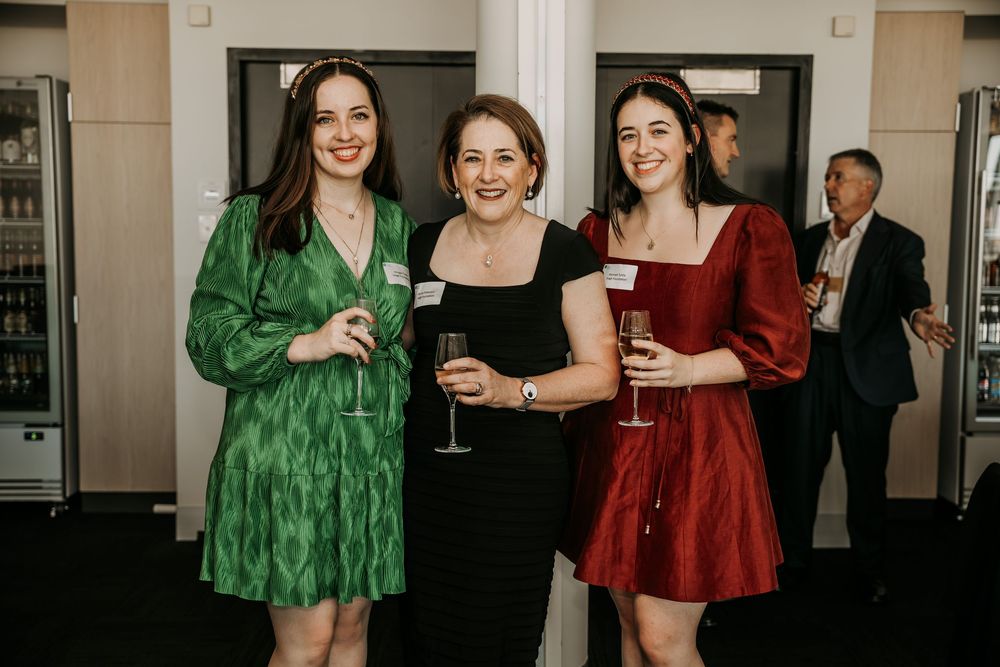 Three women in formal attire enjoying drinks at an indoor event with a partygoer visible in the background.