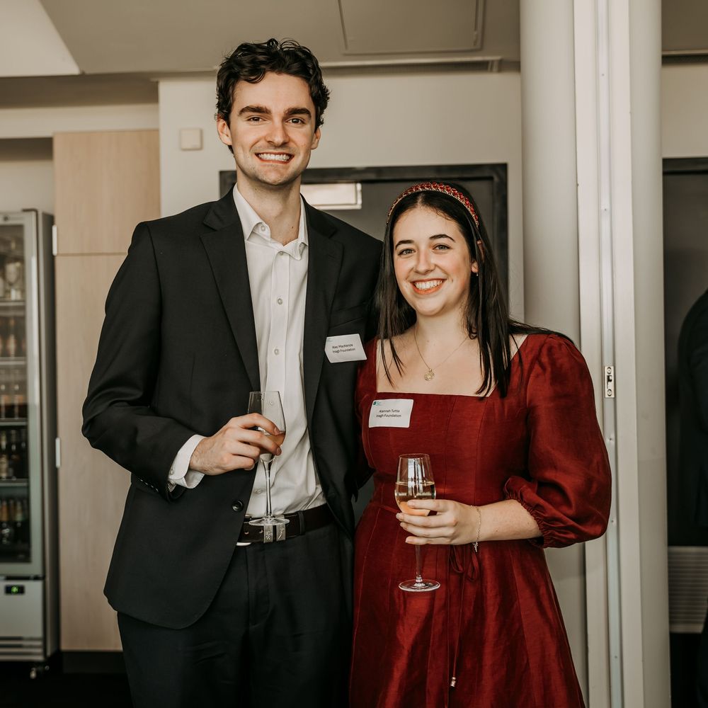 A couple in formal attire, with the woman in a burgundy dress and man in black suit, smiling at a social gathering.