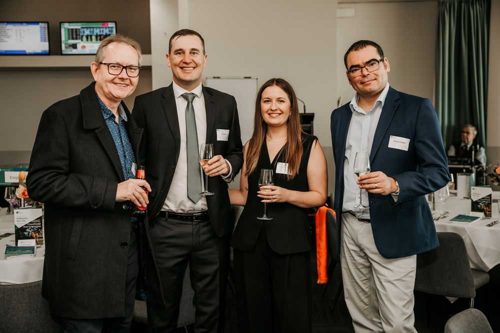 Four professionals in business attire holding drinks at a networking event in a conference room setting.