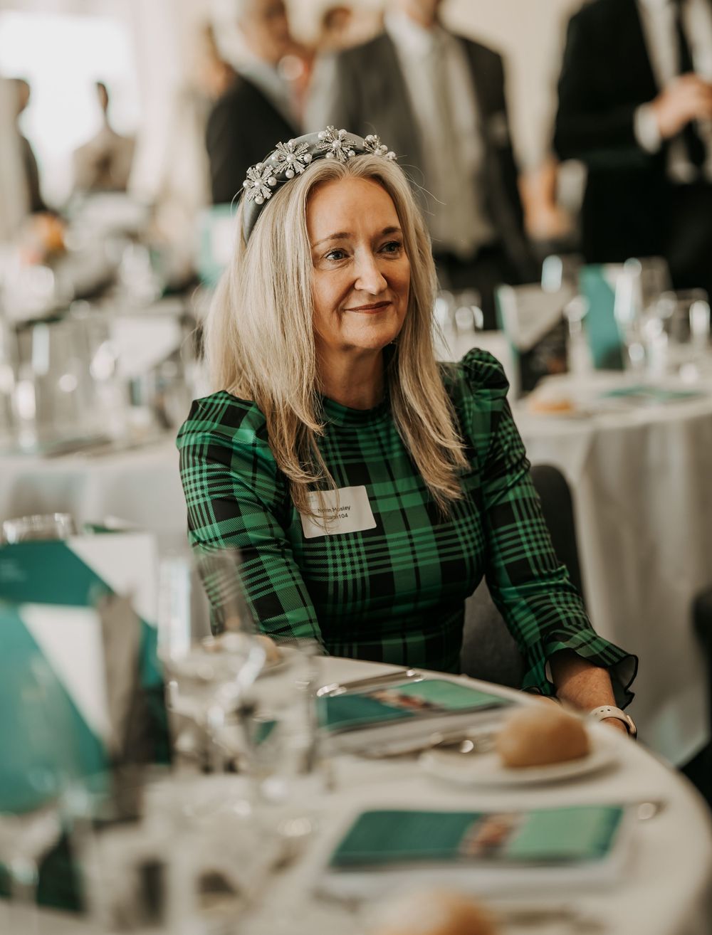 Event guest in green plaid dress with tiara sits at formal table setting with turquoise decor elements.