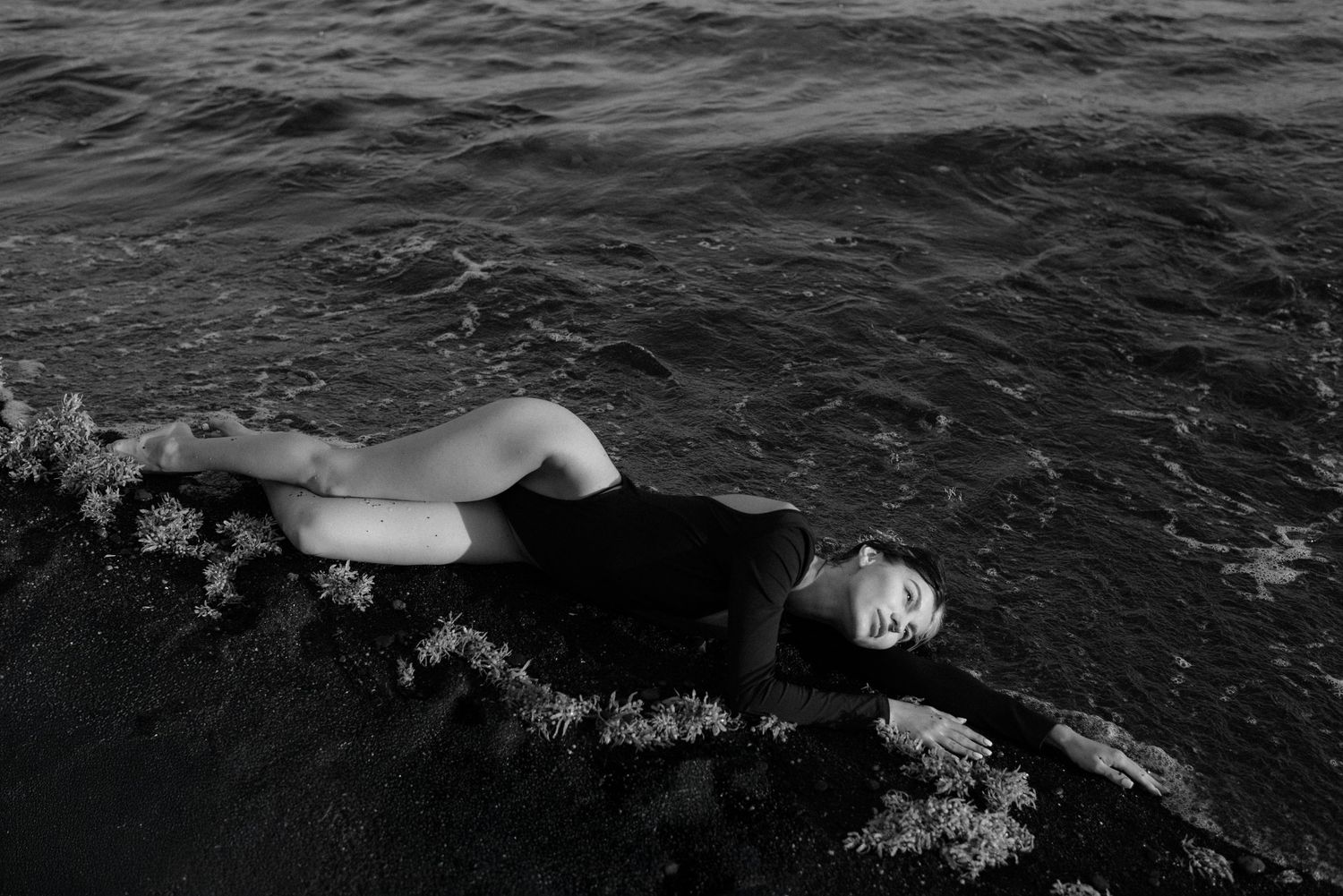 A black and white artistic photograph shows a figure floating on dark rippling water in vintage-style swimwear.