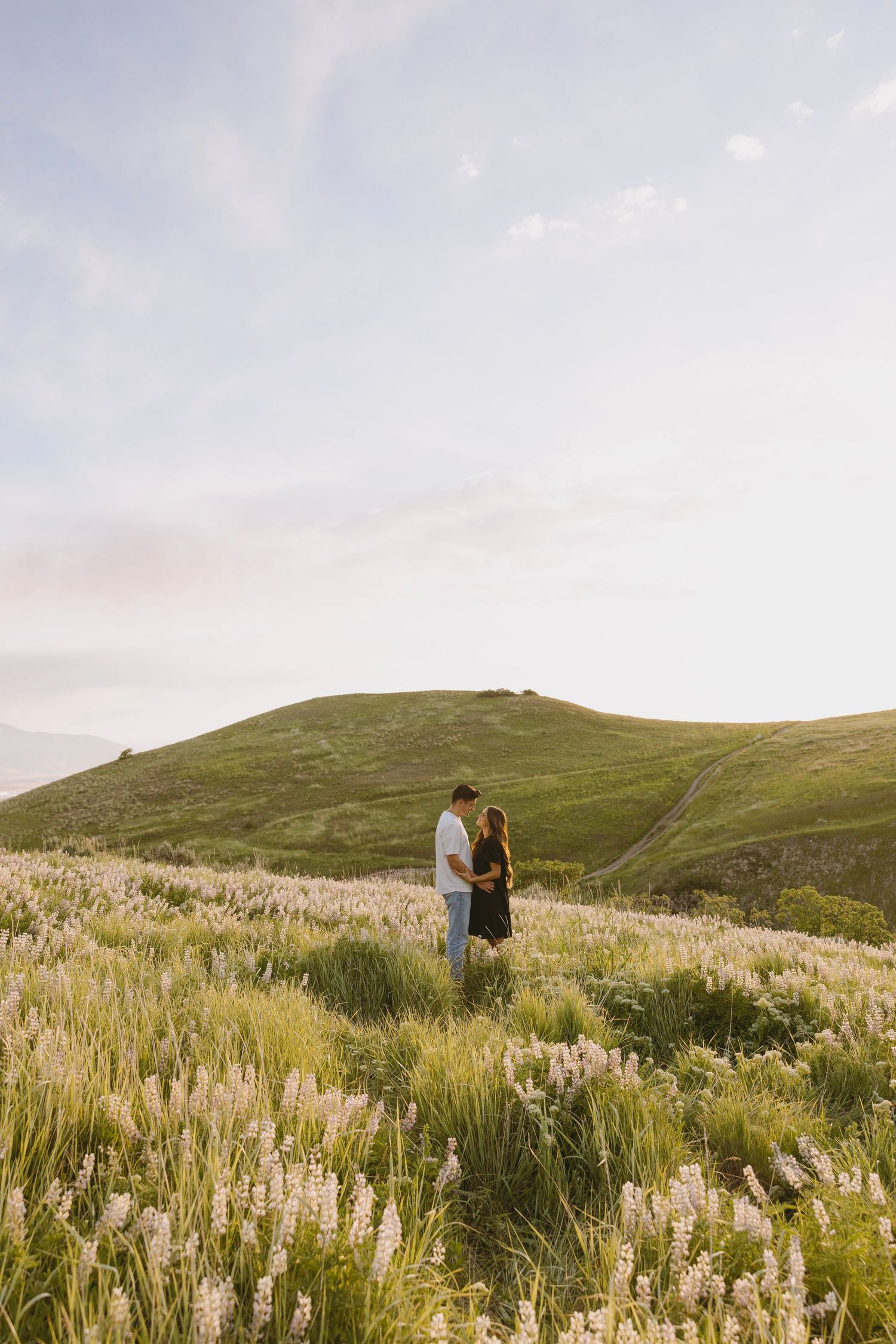Two people stand in a field of tall grass and wildflowers against rolling green hills during sunset.