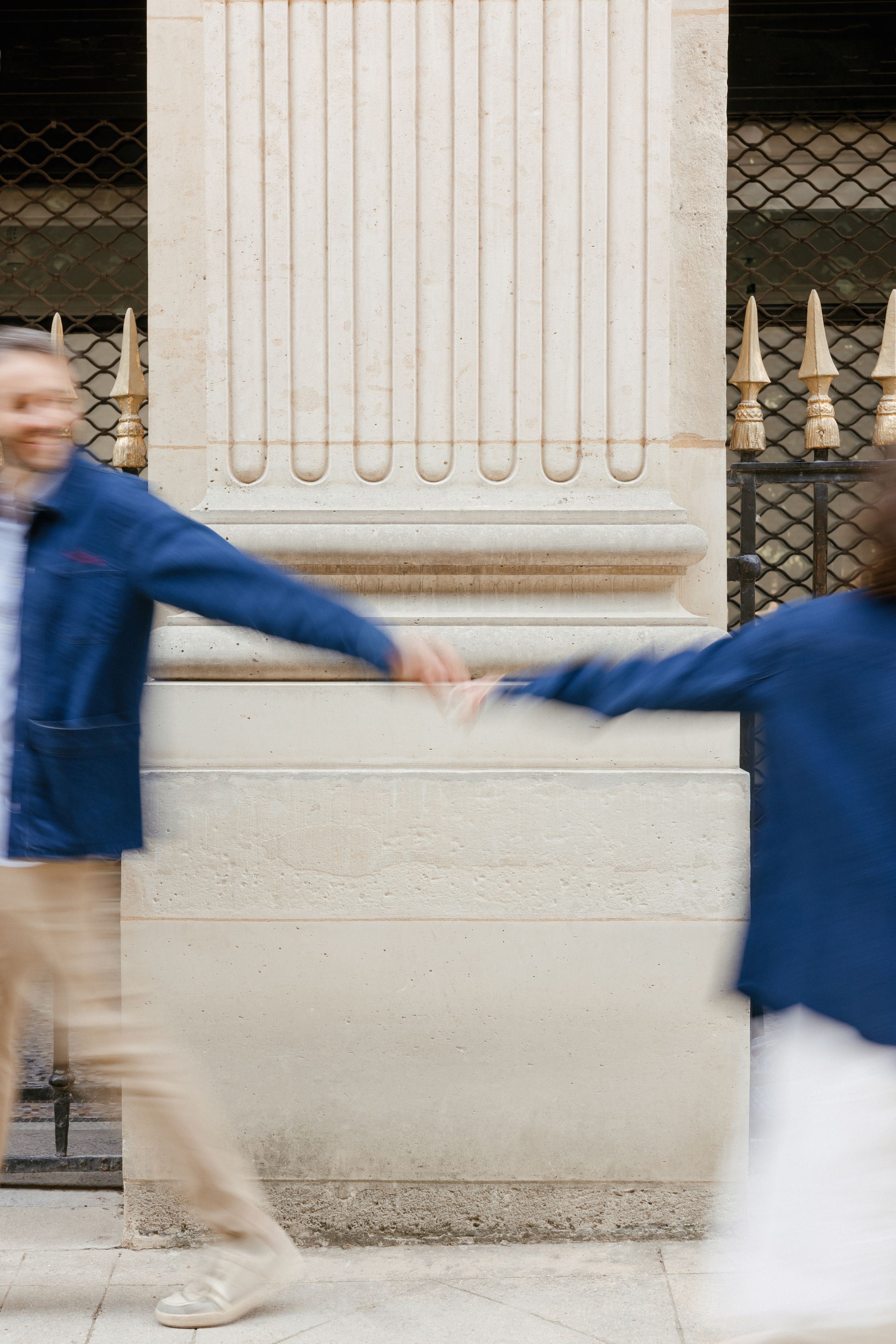 Blurred motion photo of two people in blue jackets passing each other in front of a white architectural wall.