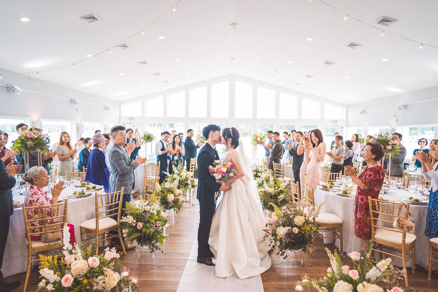 Wedding ceremony in a bright venue with floor-to-ceiling windows and elegant floral arrangements lining the aisle.