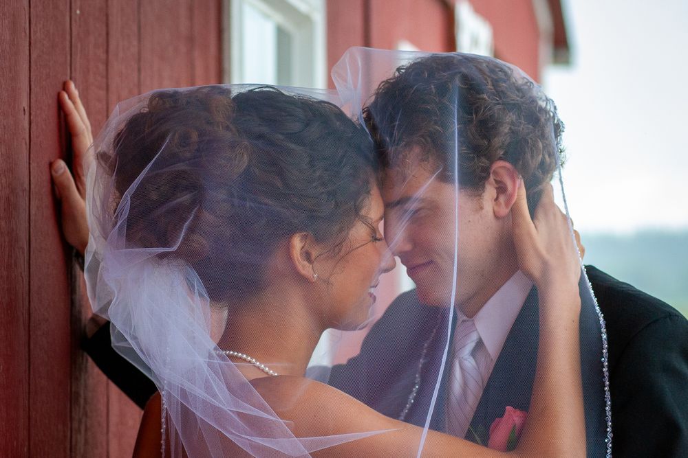 A romantic moment between newlyweds as they share a tender embrace against a red barn wall on their wedding day.