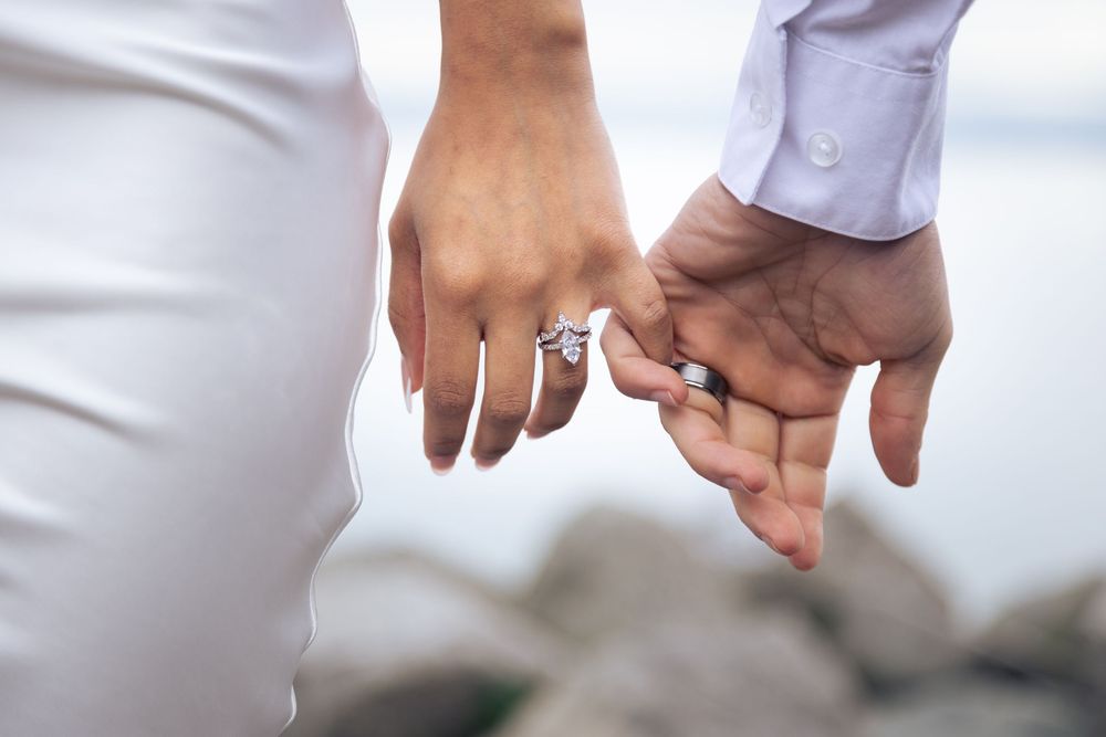 Close-up of couple holding hands with engagement ring visible, both wearing white clothing.