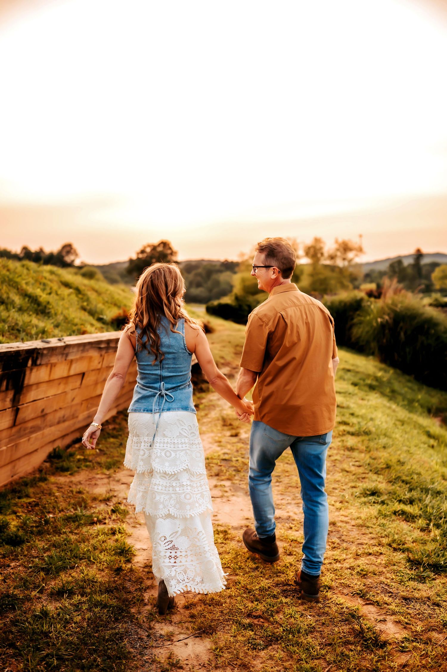 A couple walking hand in hand on a hillside path at sunset in casual denim and warm earth tones.