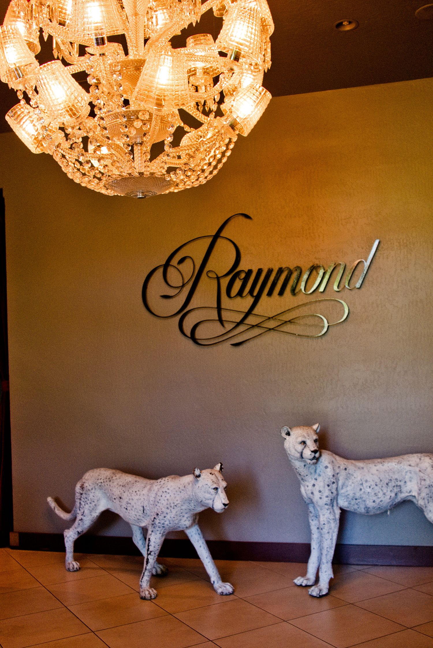 Elegant chandelier hangs above two decorative white cheetah statues against a wall with stylish 'Raymond' text signage.