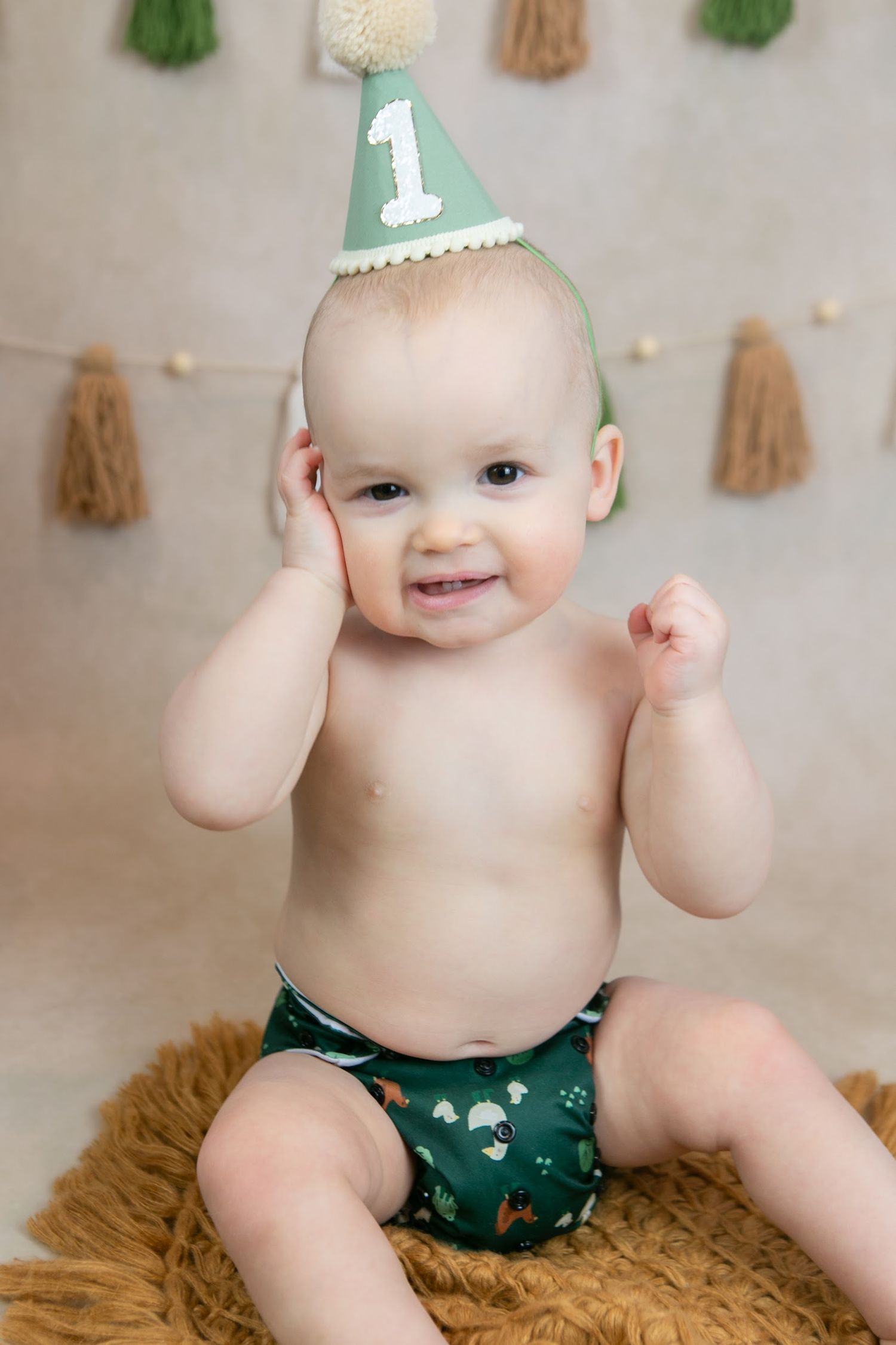 Baby in green party hat and diaper sitting on furry rug for first birthday photo shoot.