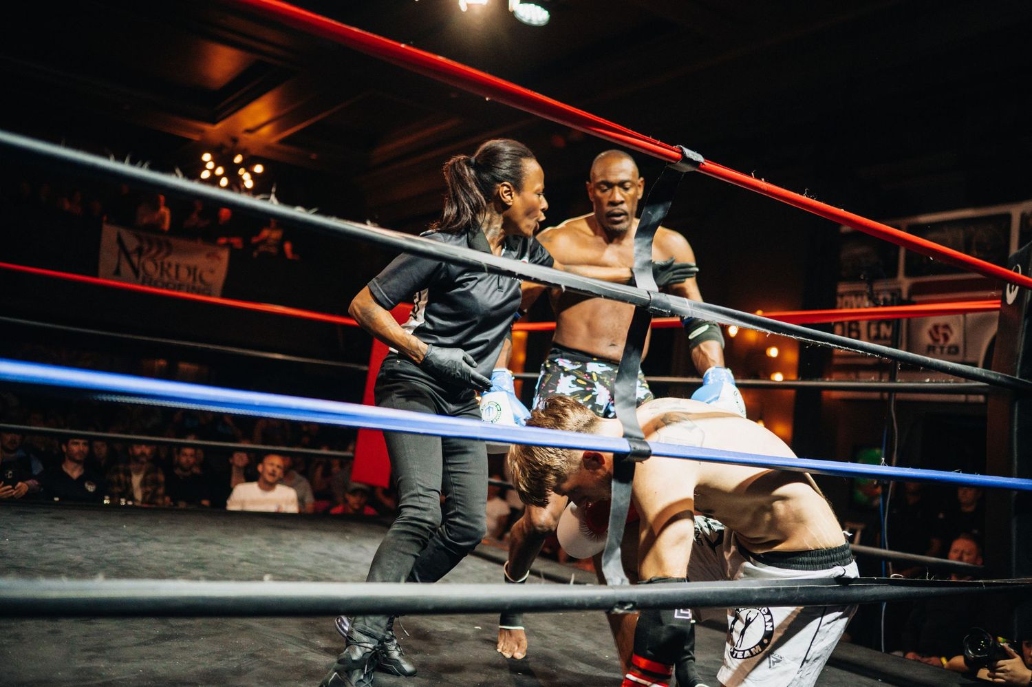 Professional wrestlers battling in a ring with ropes during an intense wrestling match under dramatic lighting.