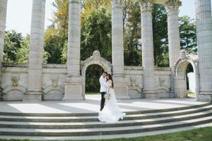 Bride and groom pose on grand stone steps surrounded by classical columns in an outdoor garden setting.