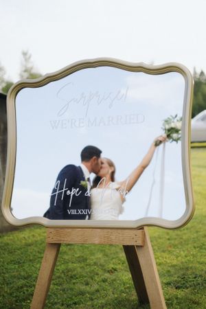 Wedding sign in ornate gold frame displayed on wooden easel in outdoor setting.