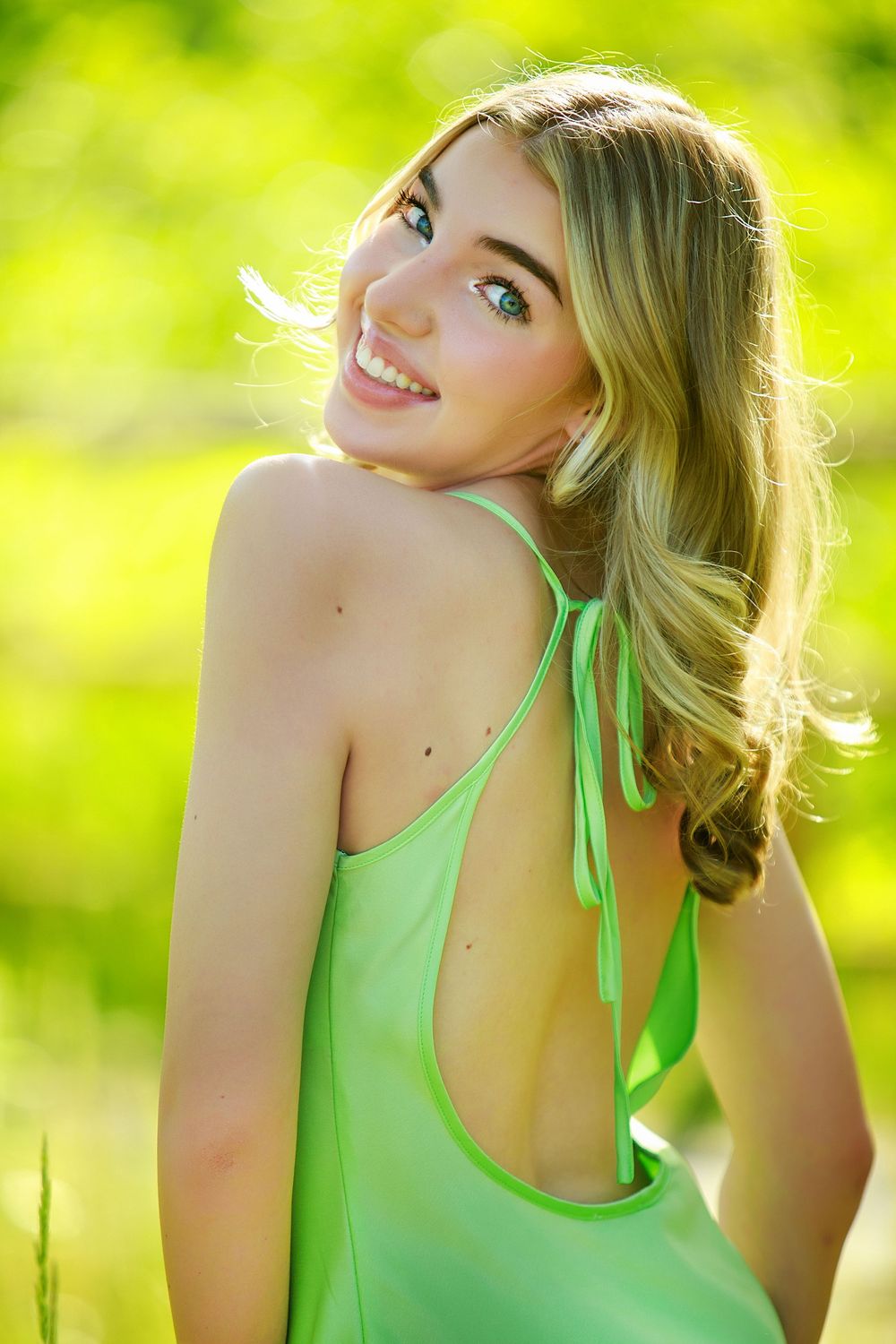 A portrait of a person with blonde hair wearing a mint green top smiling over their shoulder in bright sunlight.