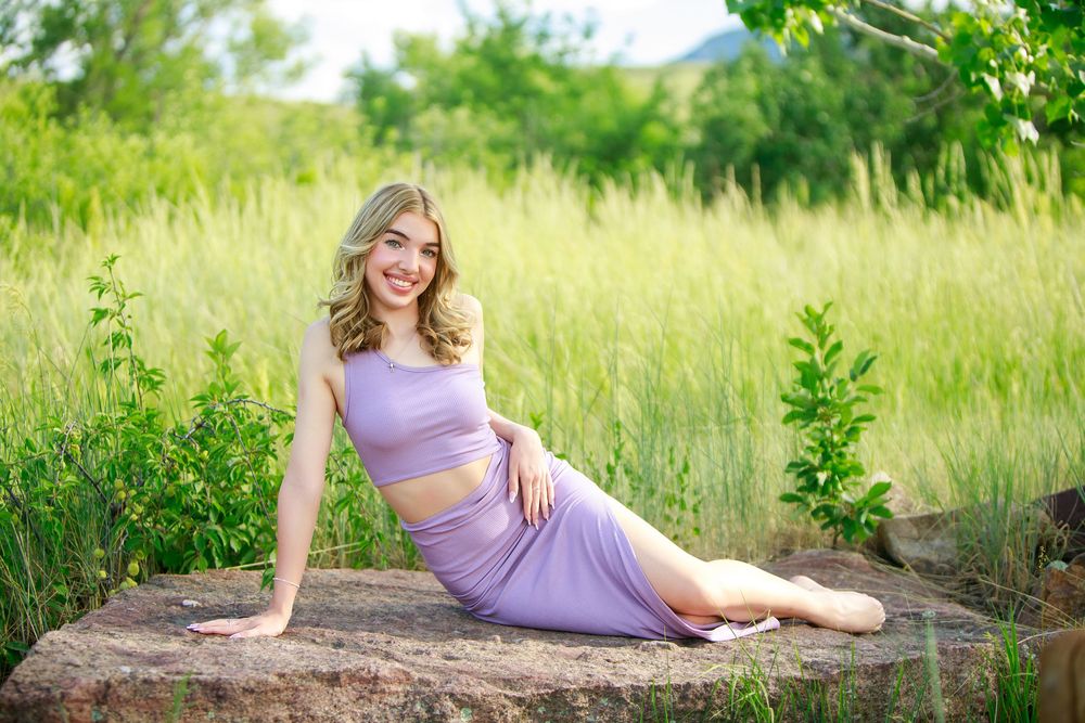 Someone in a lavender two-piece outfit poses elegantly on a rock surrounded by tall summer grass and greenery.