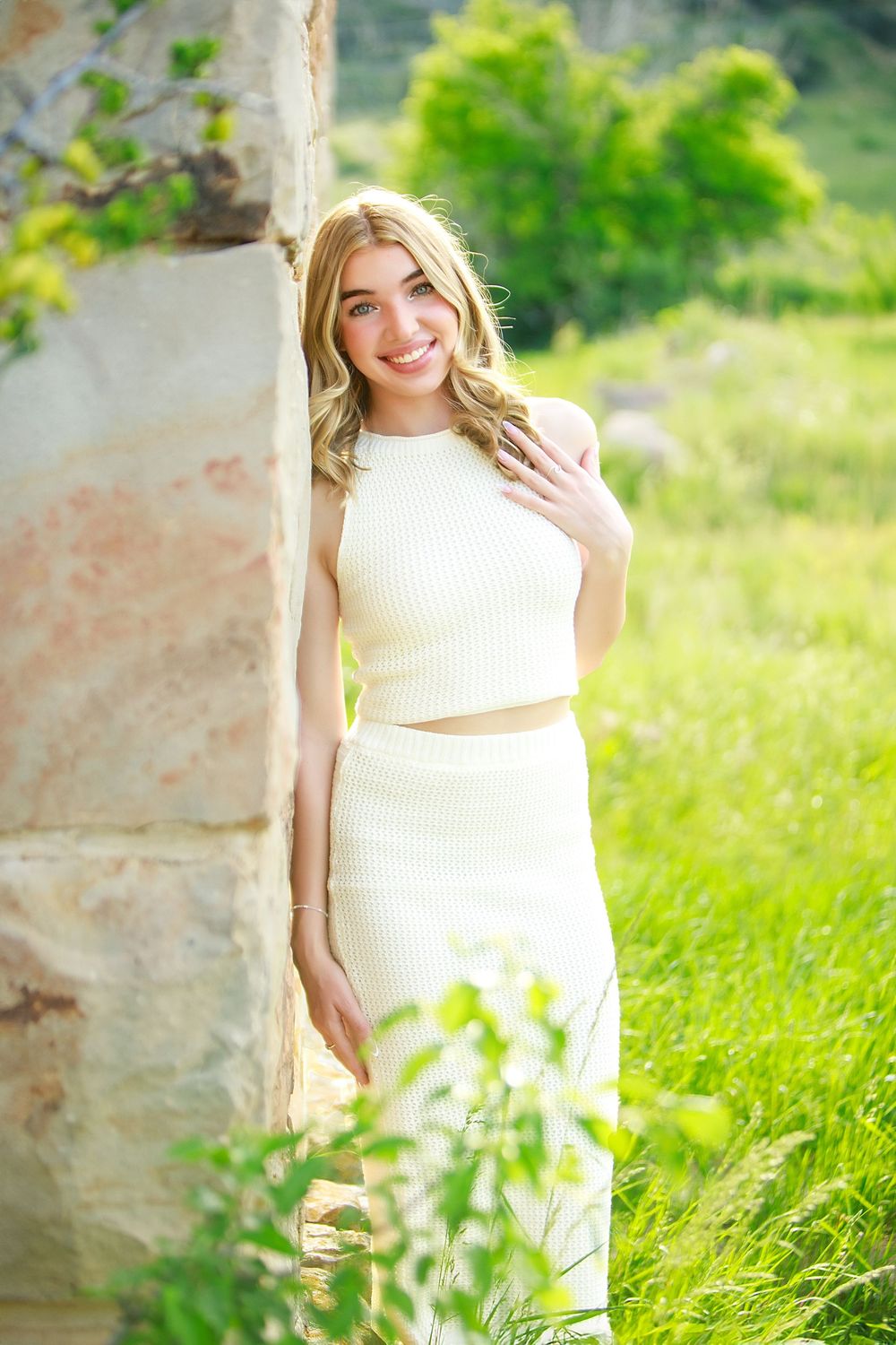 Person in a white crop top and skirt stands against stone wall outdoors in natural sunlit setting with greenery.