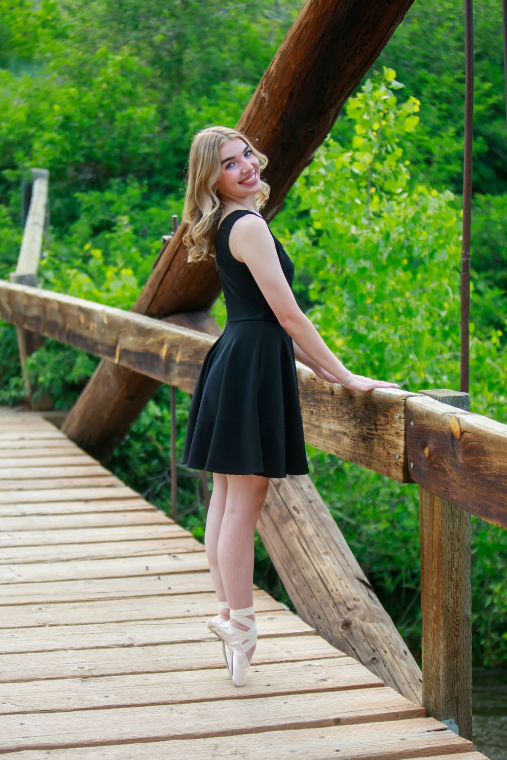 Ballet dancer in a black dress poses gracefully en pointe on a wooden bridge surrounded by lush green foliage.