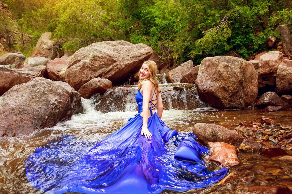 A flowing royal blue dress spreads out in a creek surrounded by large boulders and a cascading waterfall in nature.