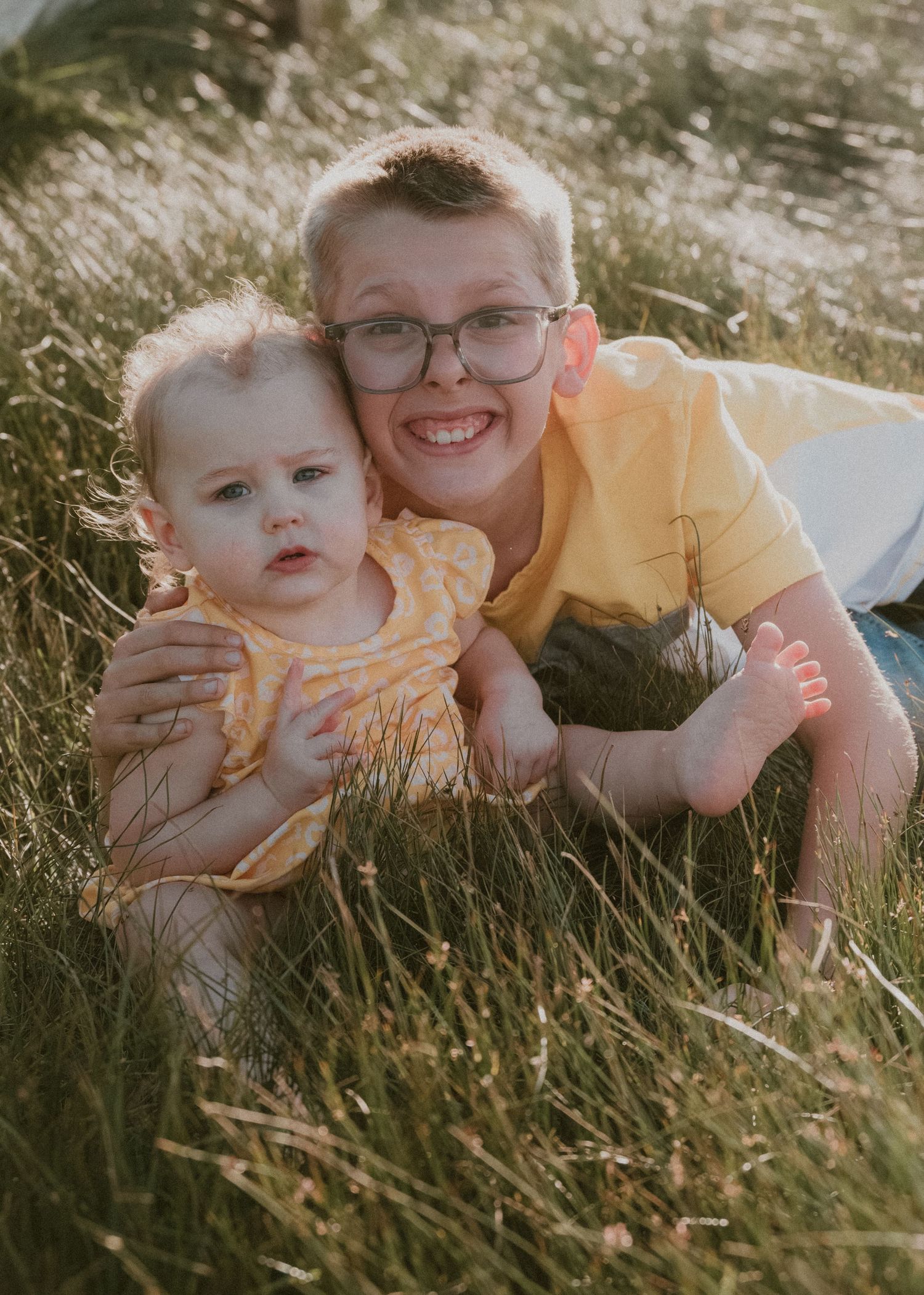 Two siblings in yellow clothing laying in tall grass during a golden hour photoshoot.