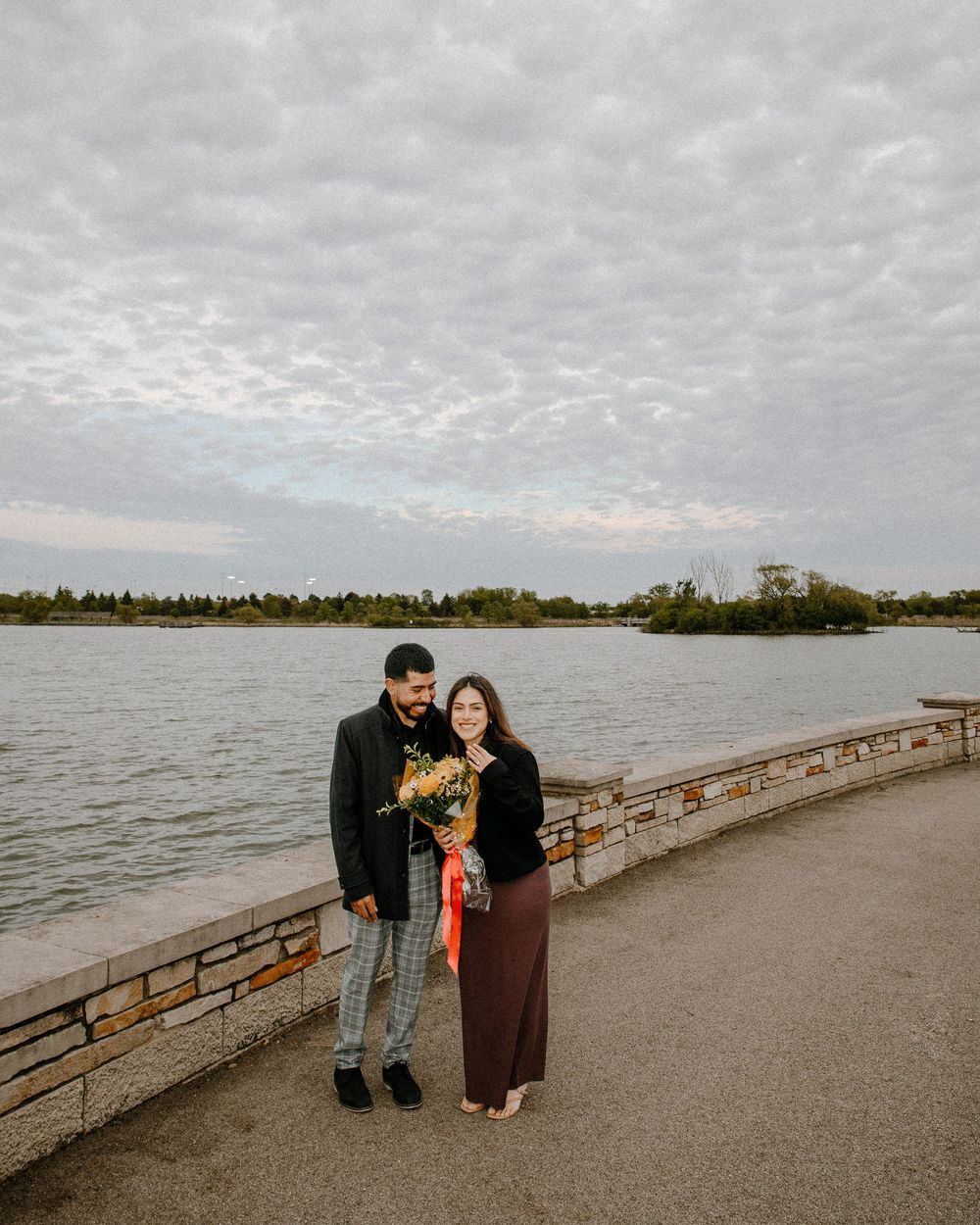 A couple stands together holding flowers on a waterfront path under cloudy skies.