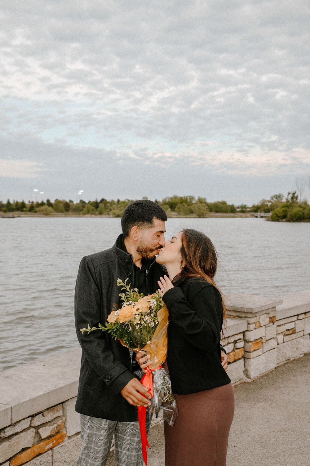 A romantic couple shares a tender kiss by a waterfront walkway while holding a small bouquet of flowers.