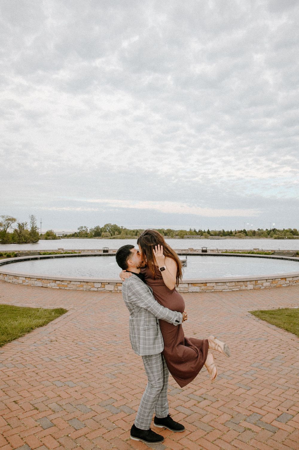 A romantic couple sharing an intimate moment on a circular brick patio overlooking a scenic lake under cloudy skies.