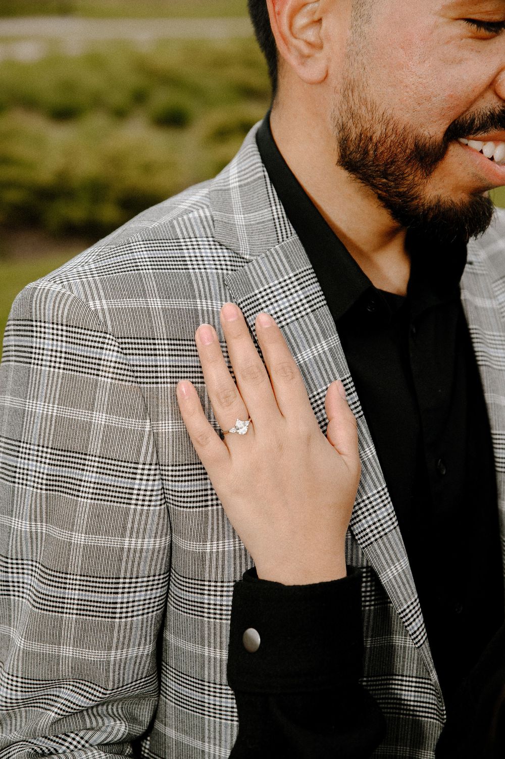 Close-up of an engagement ring displayed on a hand resting against a grey plaid suit jacket.