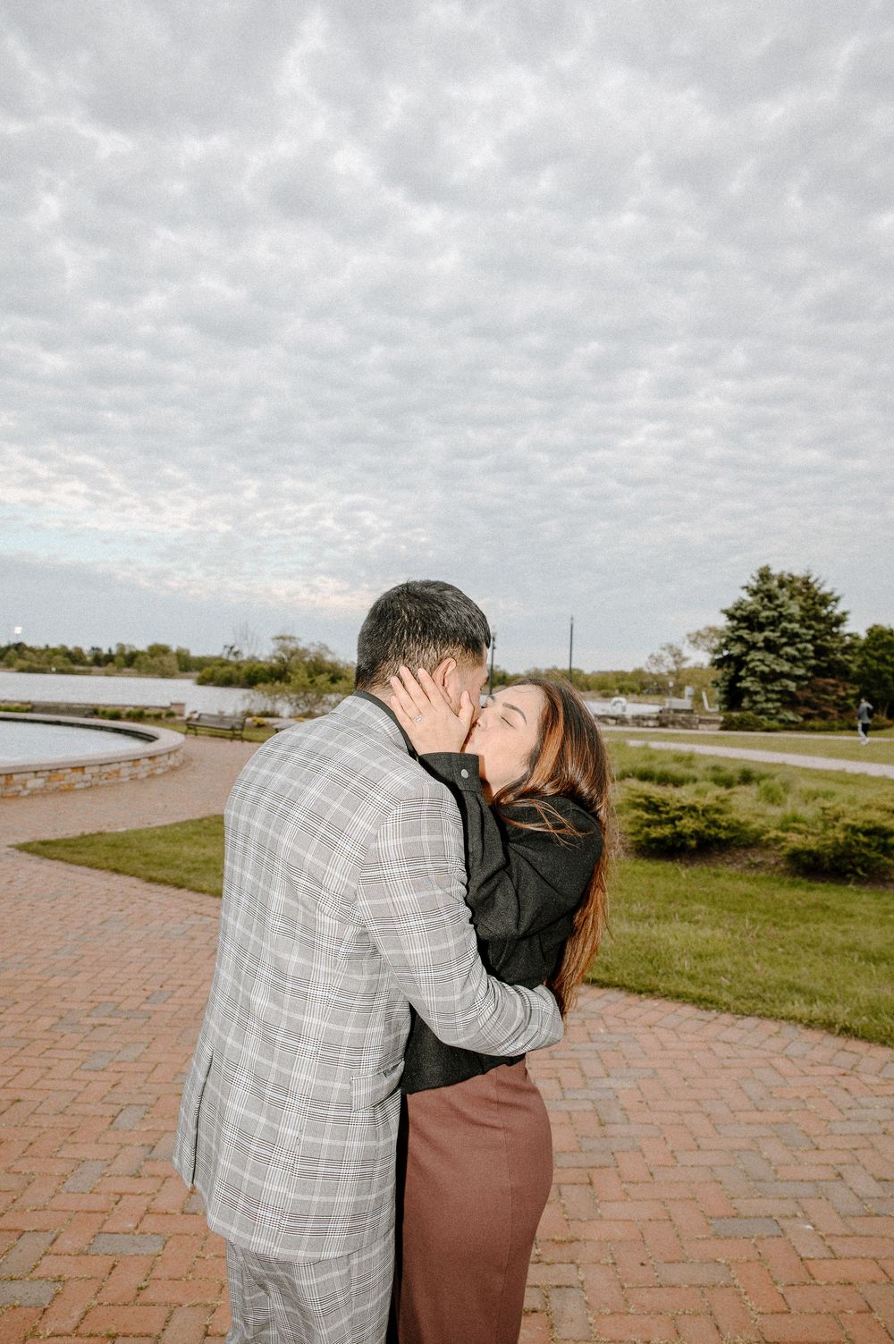 A romantic outdoor engagement moment captured against cloudy skies on a brick pathway.