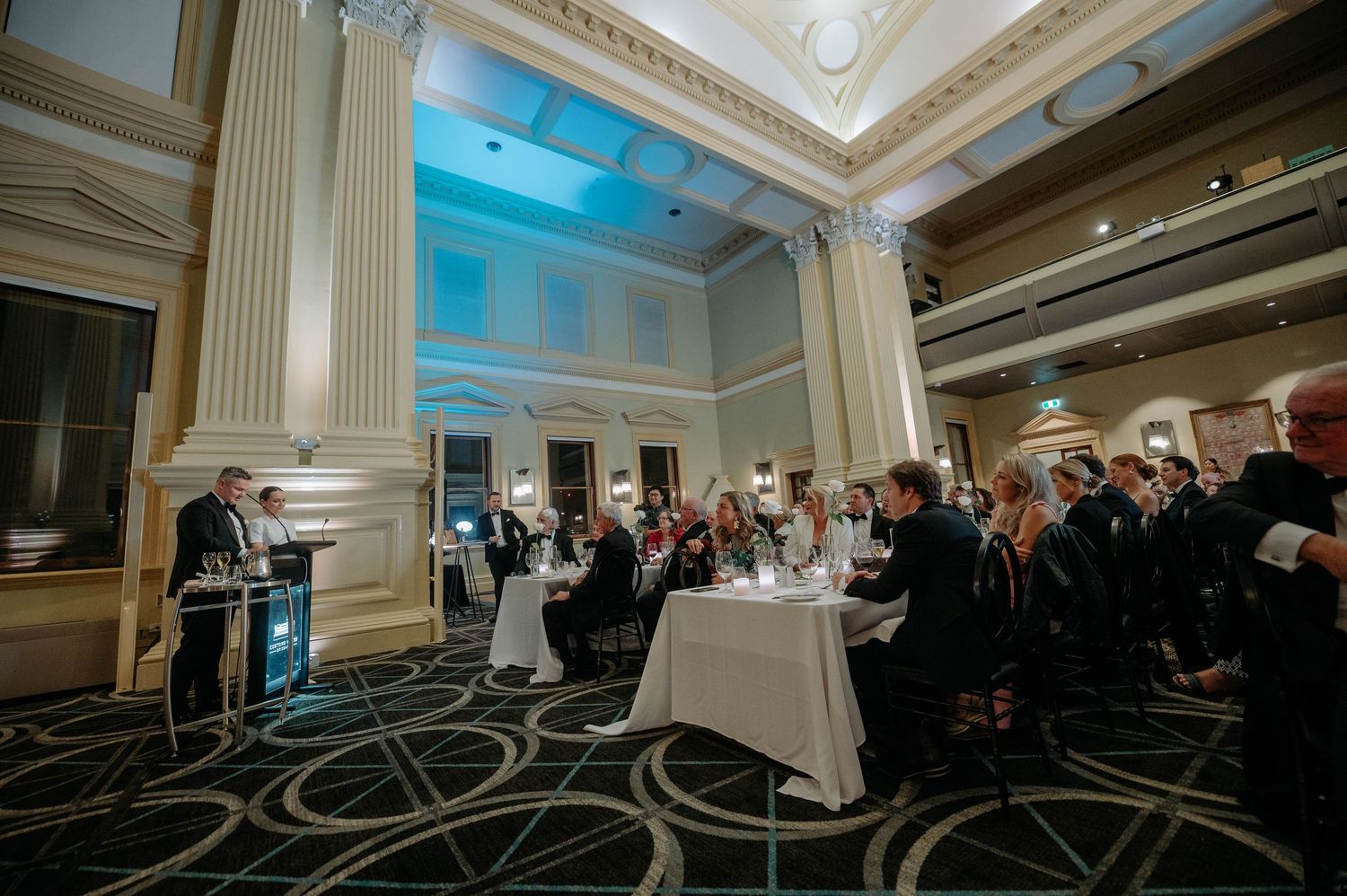 People gather in an elegant ballroom with ornate columns and a high ceiling for a formal event with round tables.