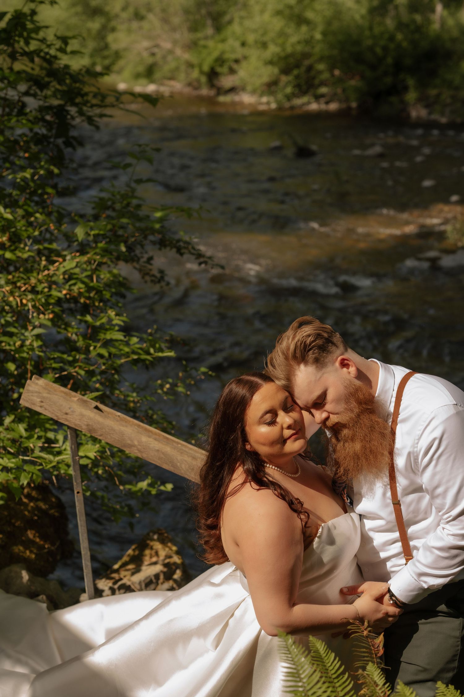 A romantic moment captured by a flowing river with natural greenery and sunlight filtering through the trees.
