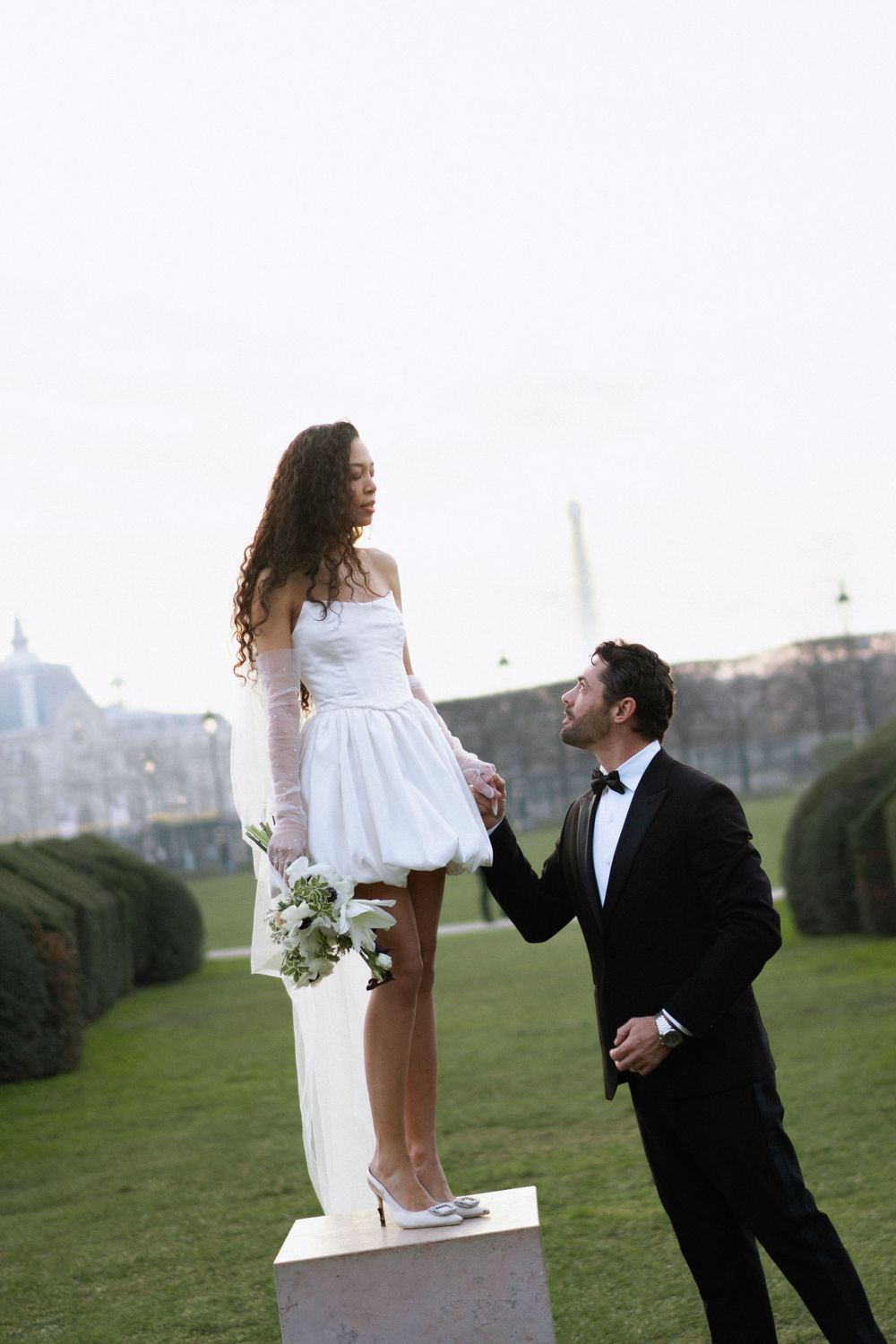 A romantic proposal scene with a bride in a short white dress standing on a pedestal in a manicured garden with Paris in view.