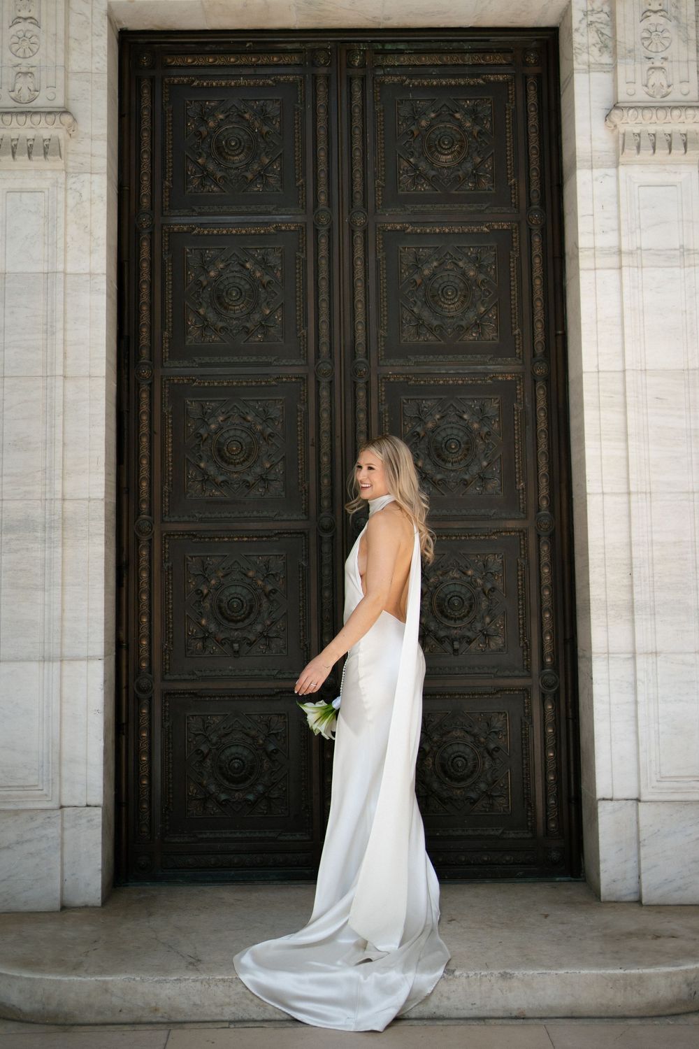 A bride in an elegant white gown stands against ornate dark carved doors in a classic architectural setting.