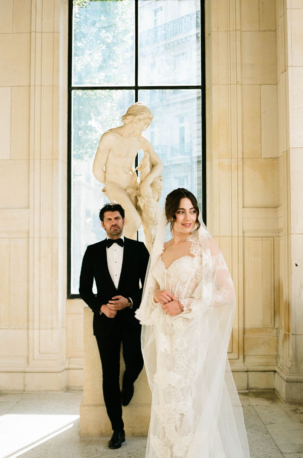 An elegant wedding couple poses in formal attire against classic stone architecture with a statue silhouetted in window light.
