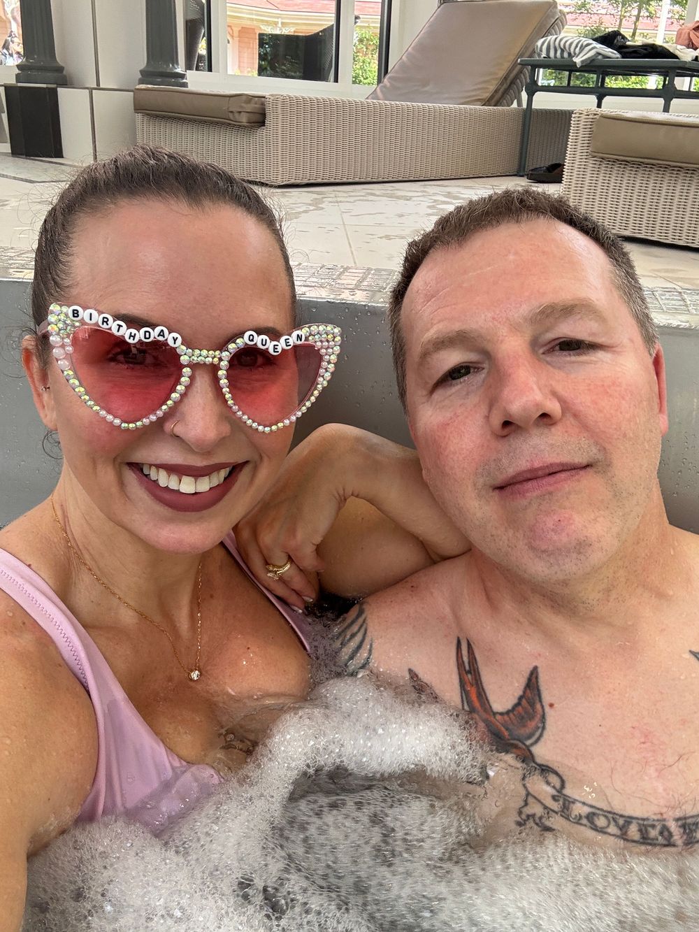 Two people smiling in a jacuzzi, with one wearing decorative heart-shaped sunglasses and the other showing visible tattoos.