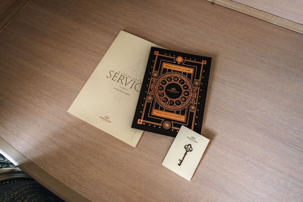 Three items laid out on a wooden desk surface: two books with ornate covers and a small card with a key design. Disneyland Paris Hotel