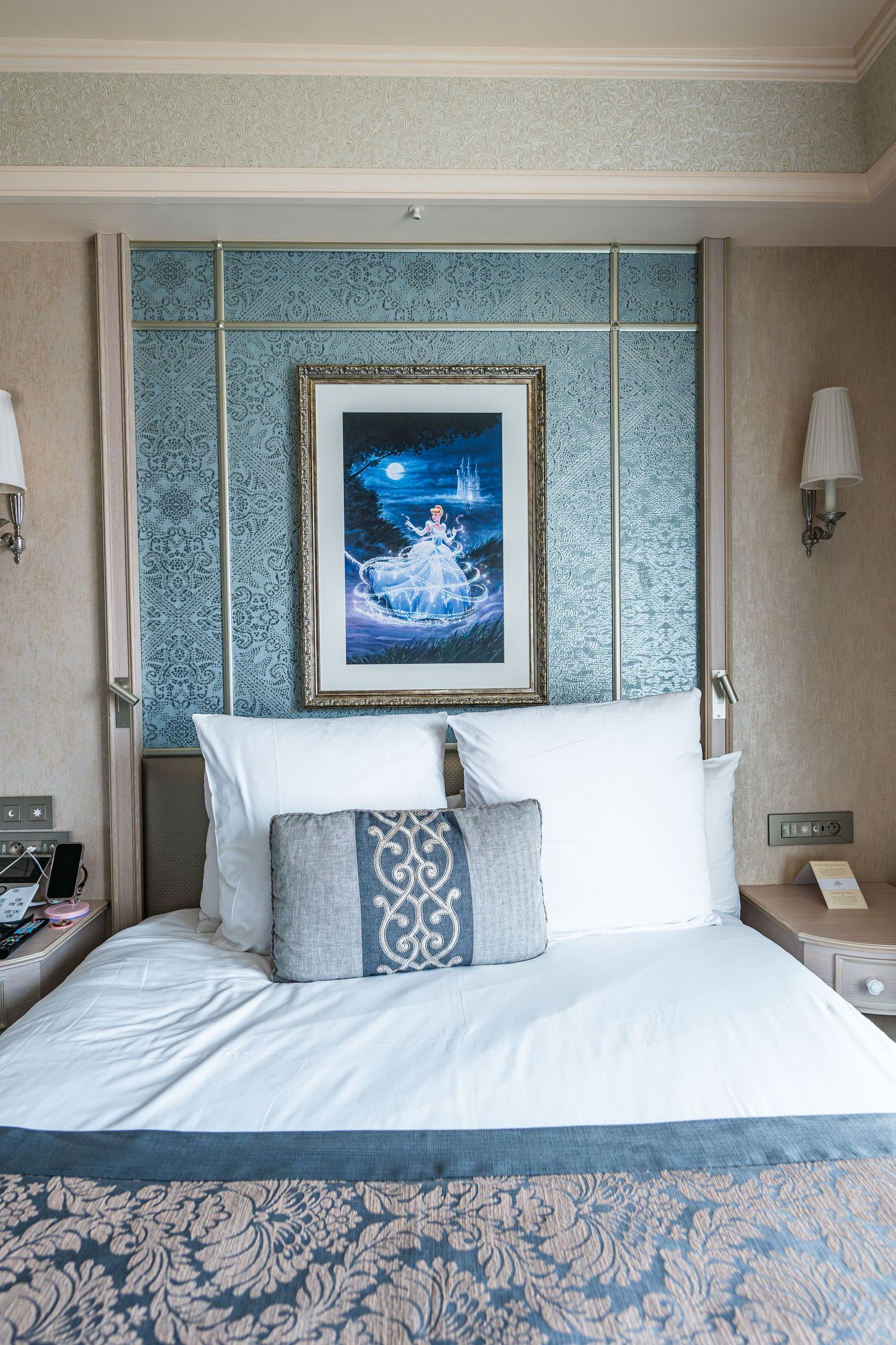 Hotel room interior with elegant blue and white decor featuring a framed artwork of Cinderella and plush bedding with damask patterns.