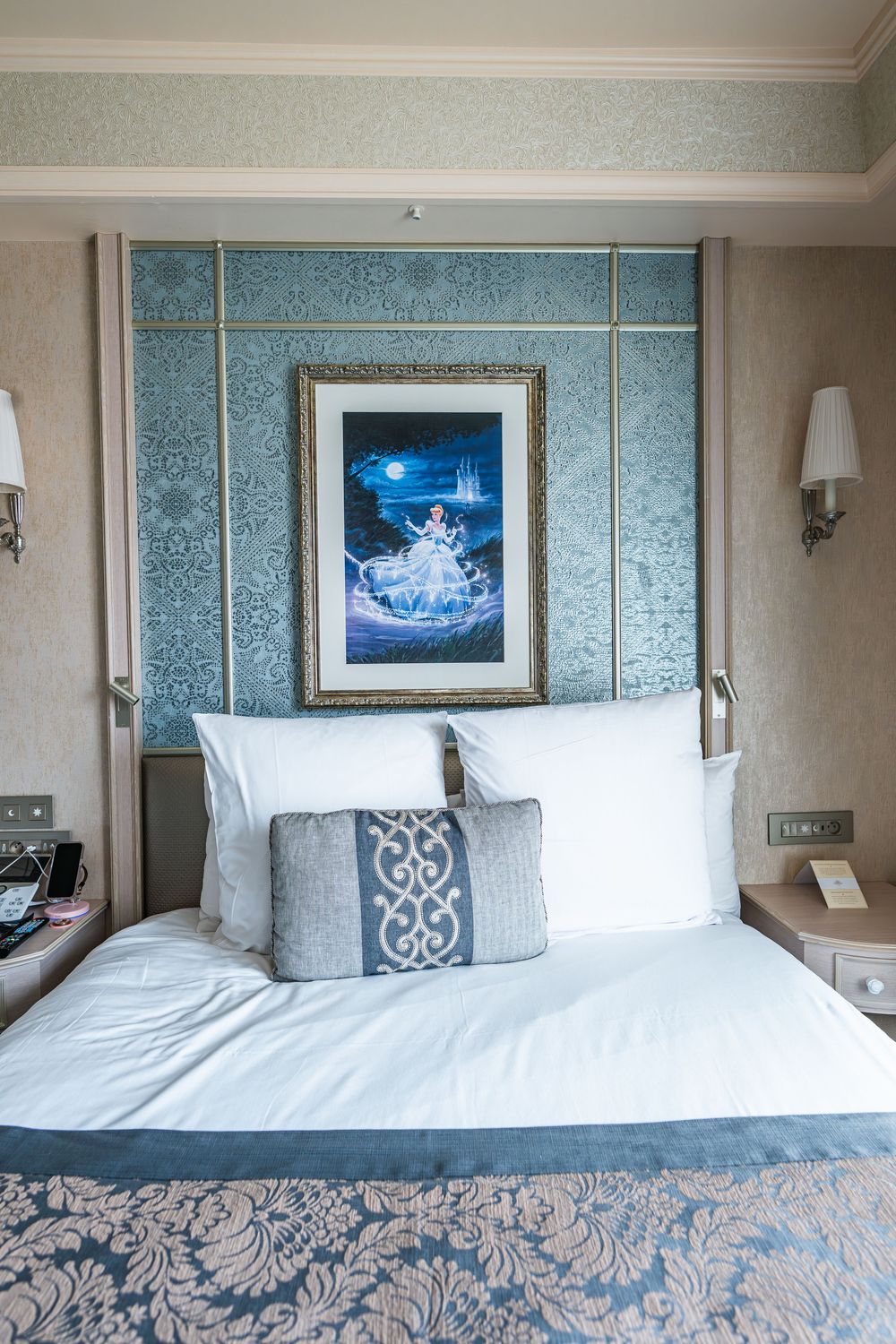 Hotel room interior with elegant blue and white decor featuring a framed artwork of Cinderella and plush bedding with damask patterns.
