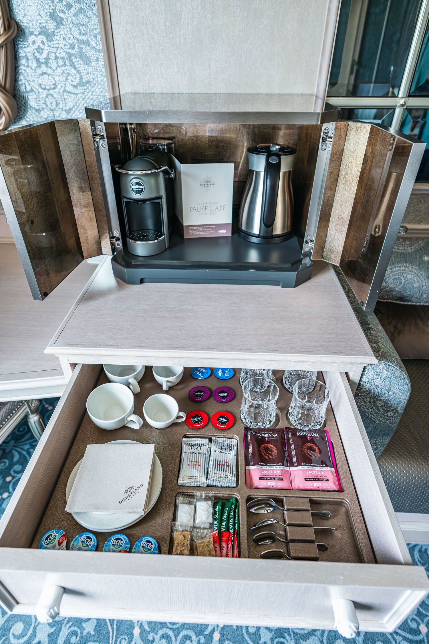 Hotel room coffee station with Keurig machine in wooden cabinet and drawer stocked with coffee pods and supplies.