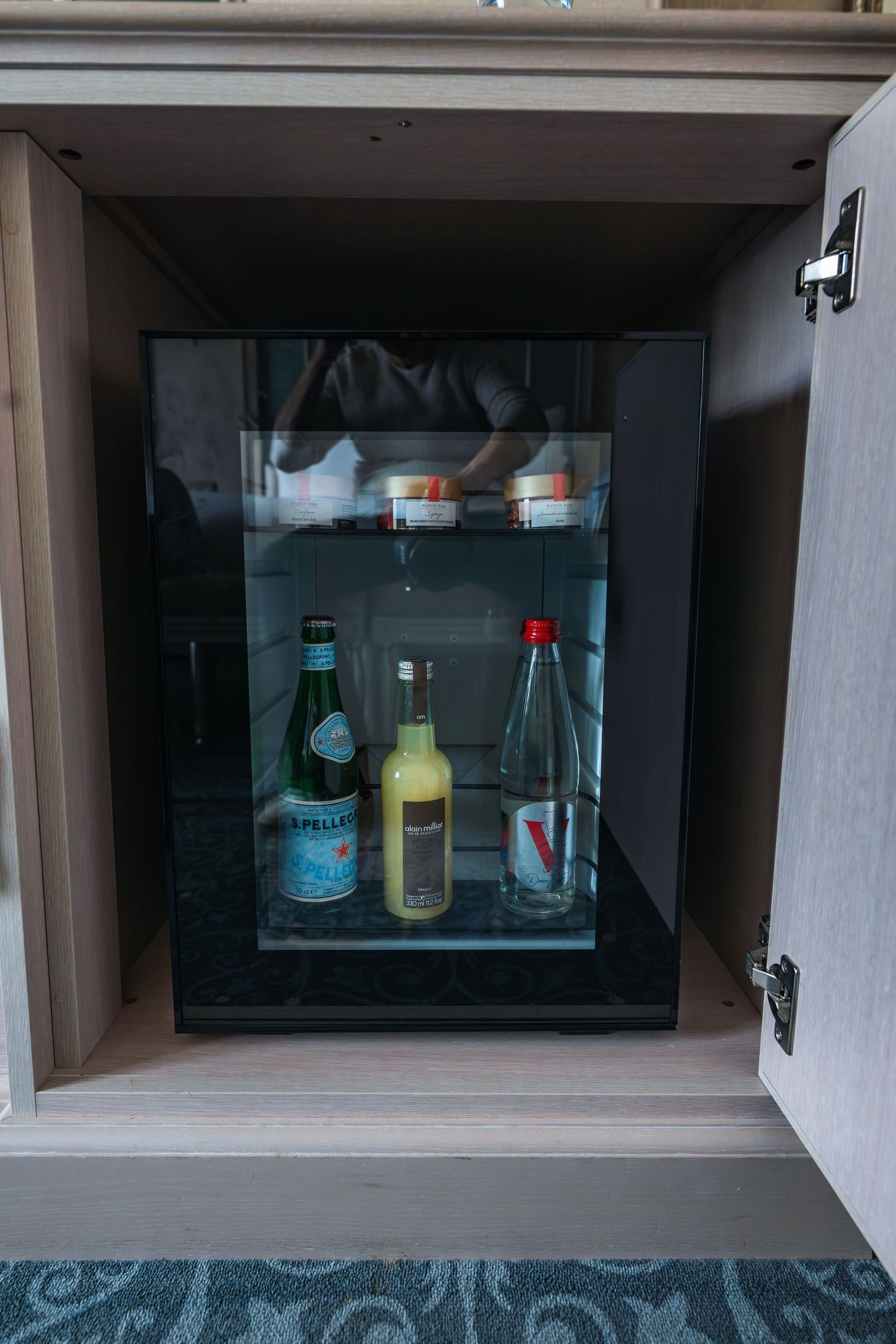 A small mini fridge with glass door displaying various bottled beverages and drinks inside a cabinet compartment.