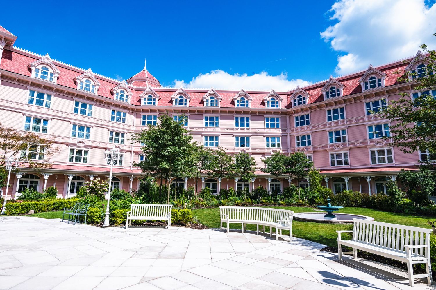 Pink-hued luxury hotel building exterior with manicured courtyard gardens and white benches under bright blue sky.