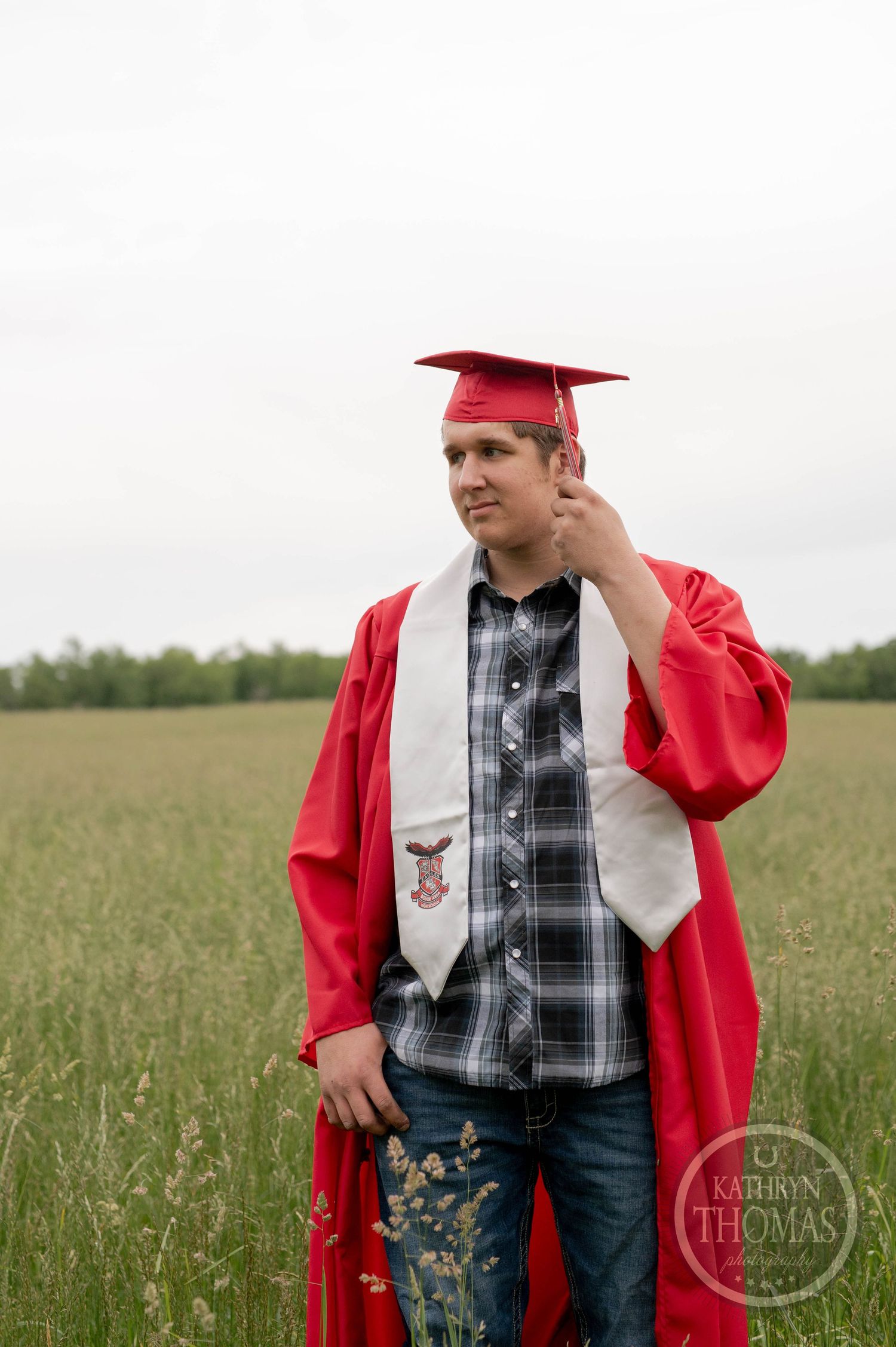 Graduate in red cap and gown stands in a grassy field wearing plaid shirt and graduation stole.