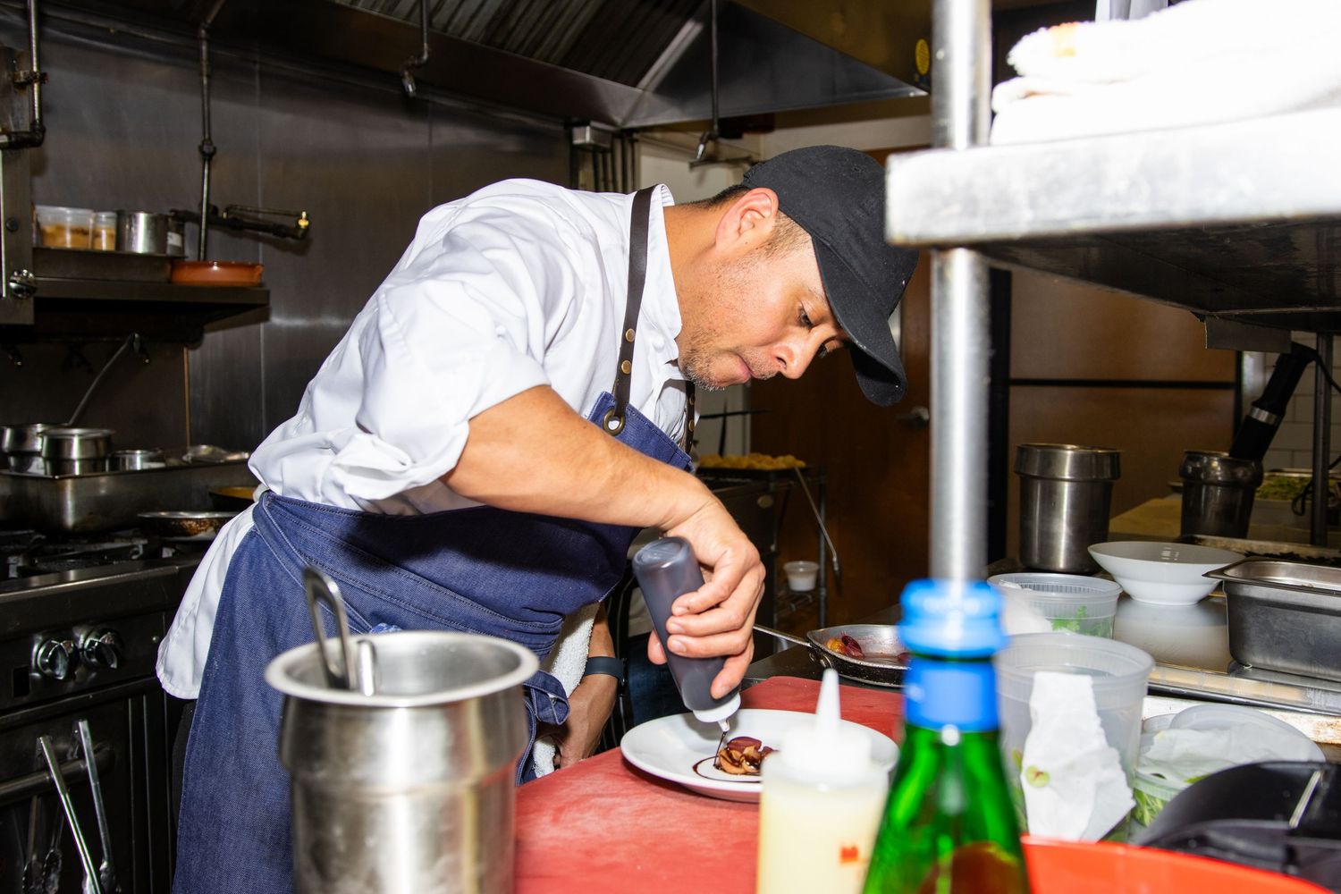 Chef in white shirt and blue apron carefully seasons food in a metal pot at commercial kitchen workstation.