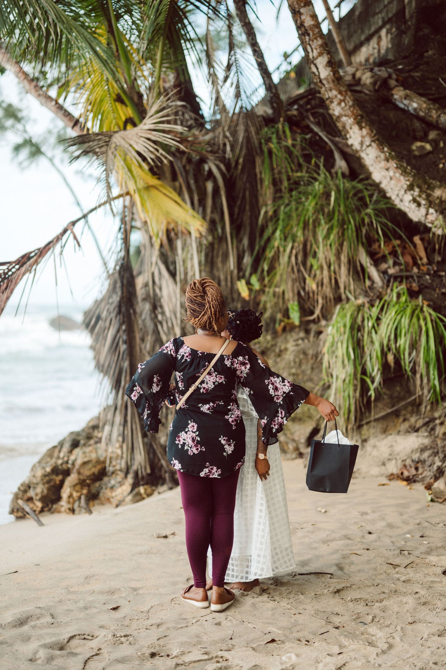 Two figures walk along a tropical beach with palm trees and rocky cliffs, carrying a white bag on a cloudy day.