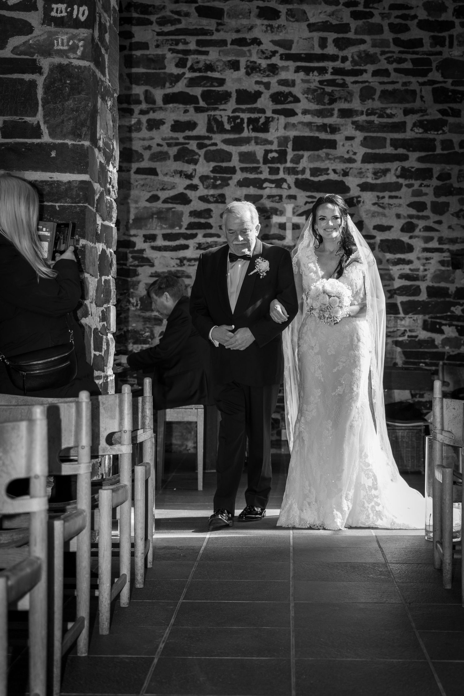 Bride in lace gown walks down stone church aisle with older gentleman in tuxedo during wedding ceremony.