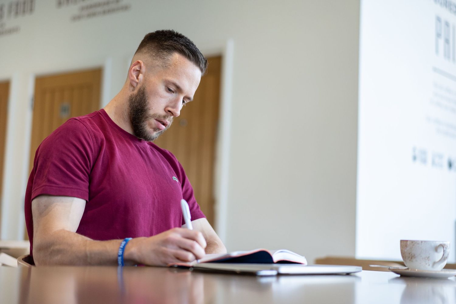 Successful business person in a burgundy t-shirt sits at a table writing in a notebook with a cup of coffee nearby.