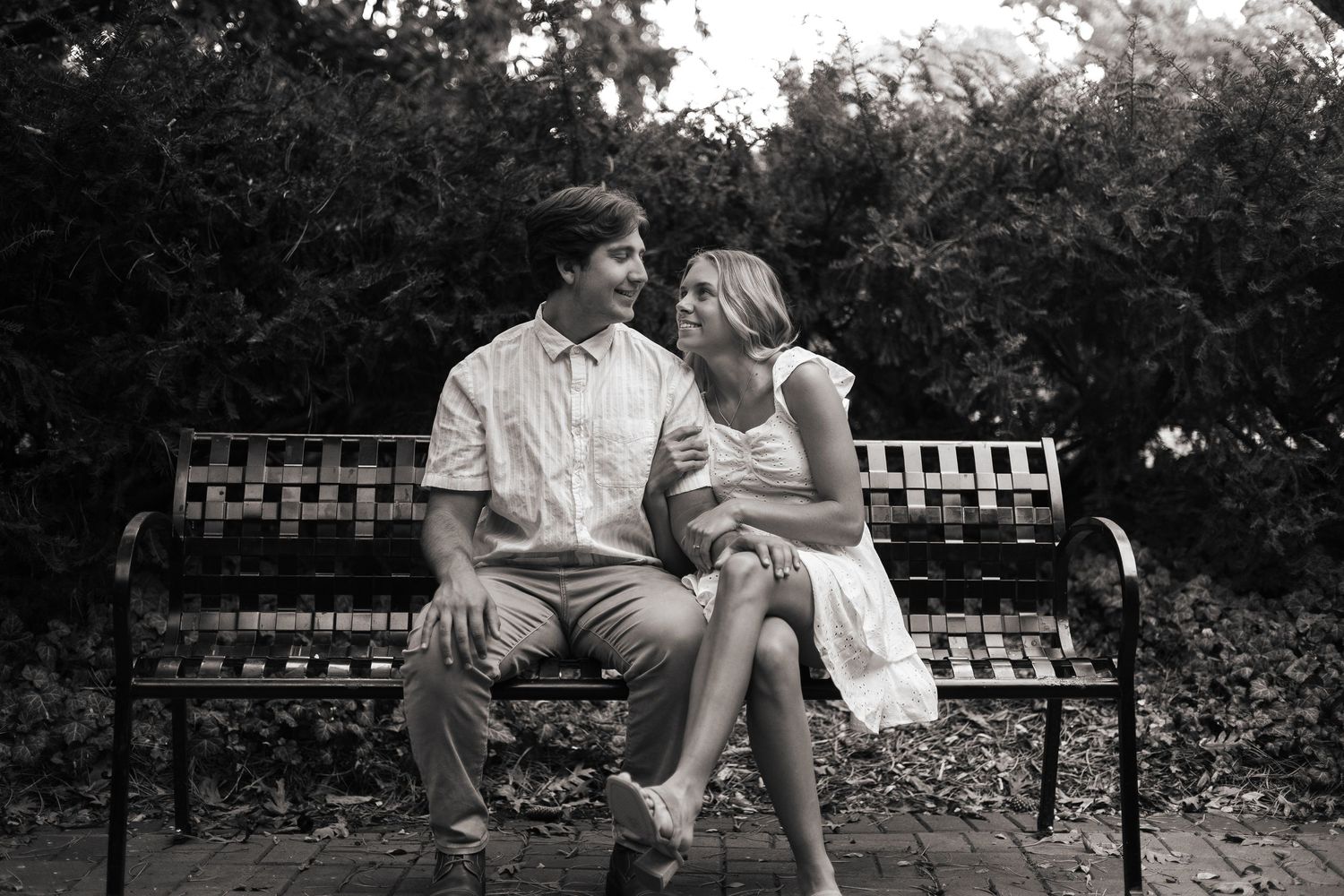 A couple sits close together on a park bench surrounded by lush greenery and trees.