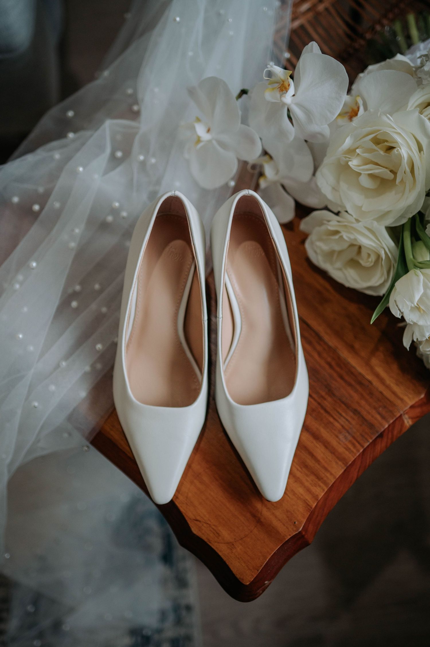 Elegant white bridal heels displayed with white roses and delicate veil on wooden surface.