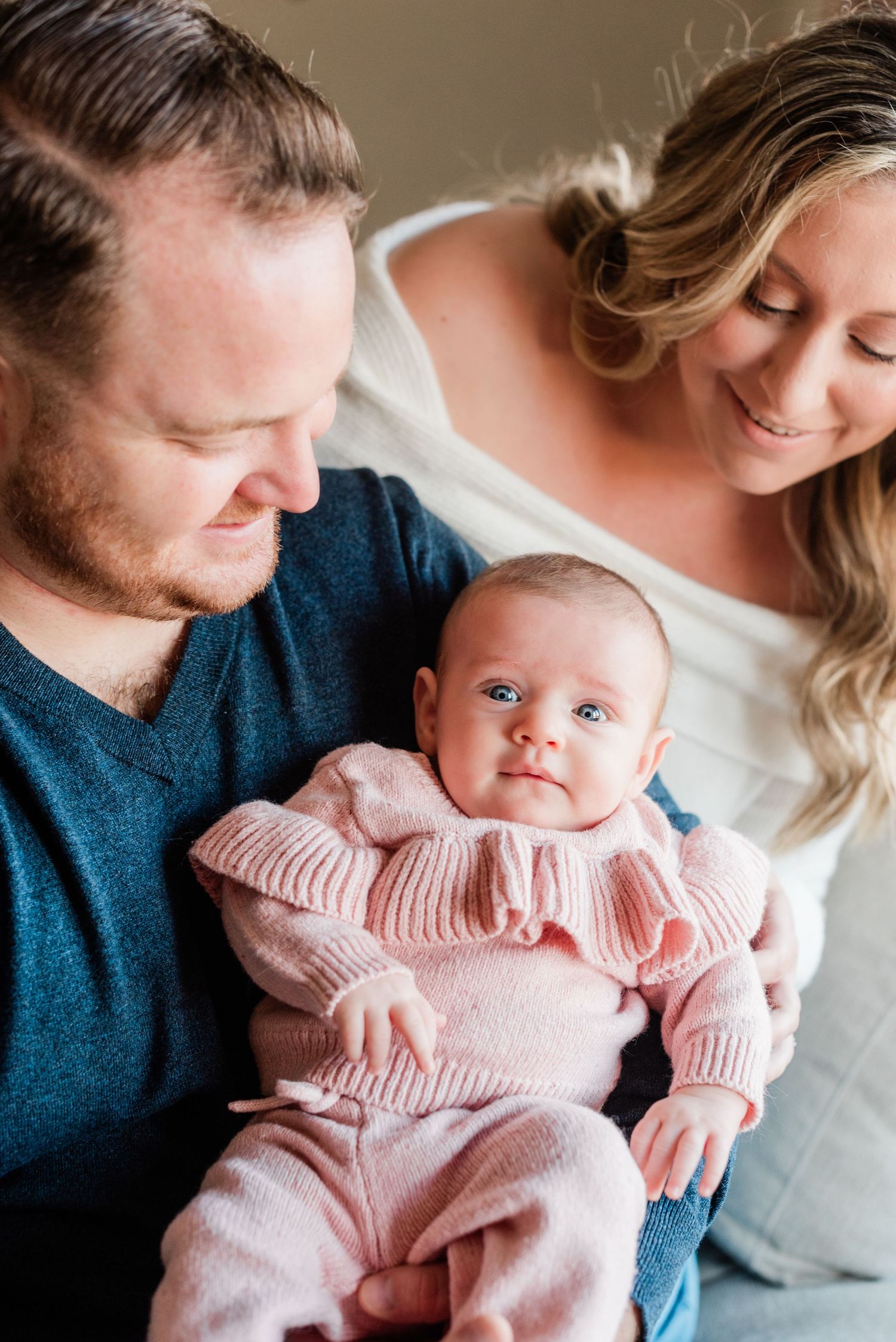 A couple shares an intimate moment with their newborn baby wearing a pink sweater while relaxing at home.