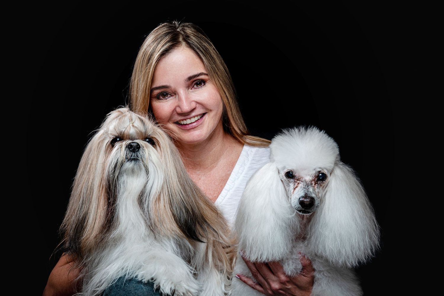 A person in a white shirt poses with two fluffy white Shih Tzu dogs against a dark background.